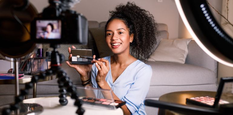 A content creator filming a makeup tutorial at home. She is holding a palette and smiling at the camera, surrounded by professional lighting and makeup products, exemplifying content marketing in the beauty industry.