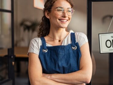 A cheerful girl with specs folded hands, blue apron infront of a store