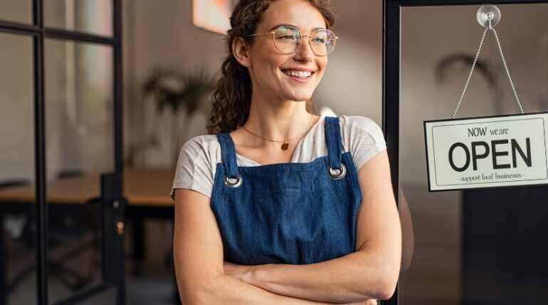 A cheerful girl with specs folded hands, blue apron infront of a store